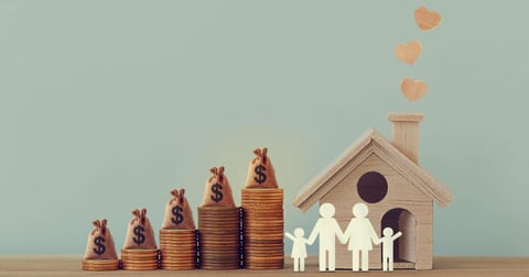 Mini house and heart, family members, US money bags on rows of rising coins on a table.